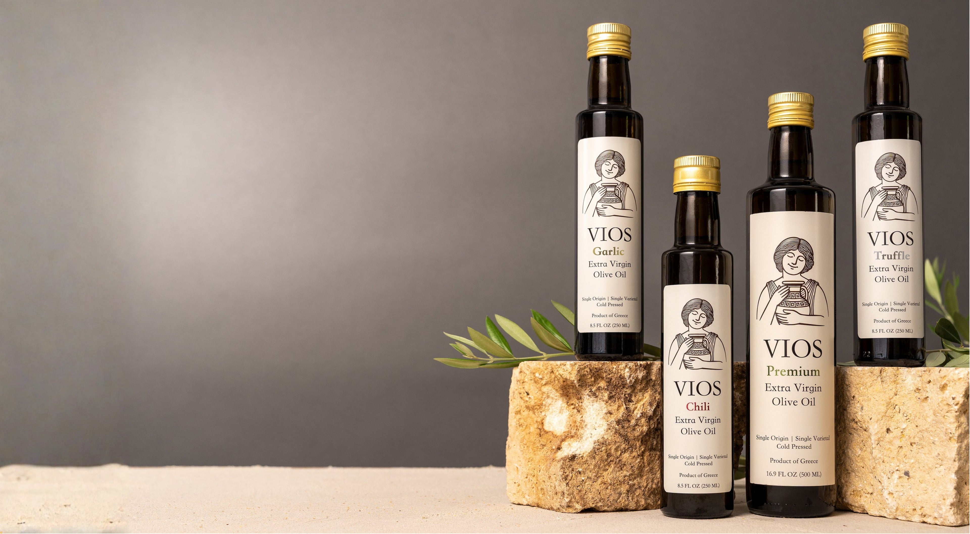 Four bottles of VIOS olive oil on a stone surface with olive branches against a gray background