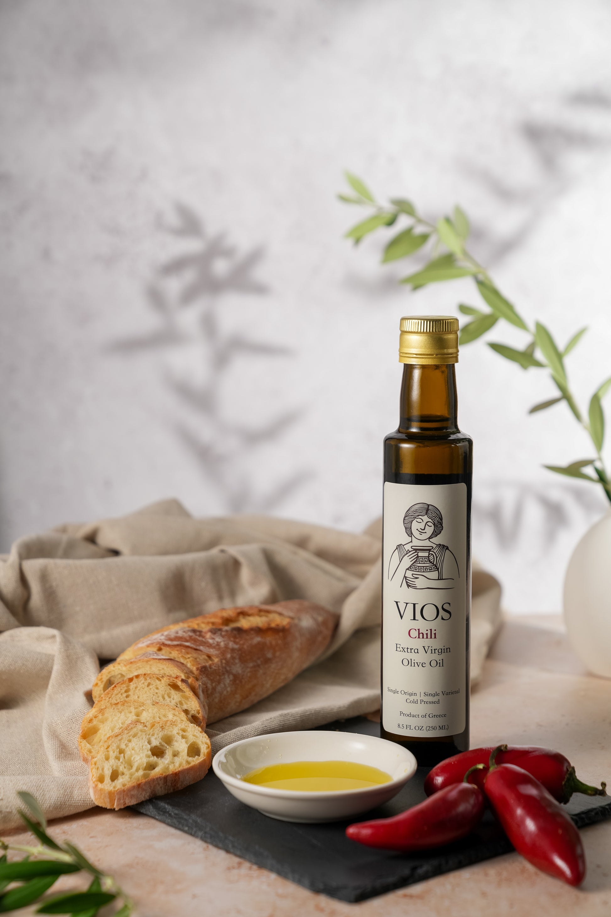Bottle of VIOS chili extra virgin olive oil with bread, red chili peppers, and a saucer filled with olive oil on a table
