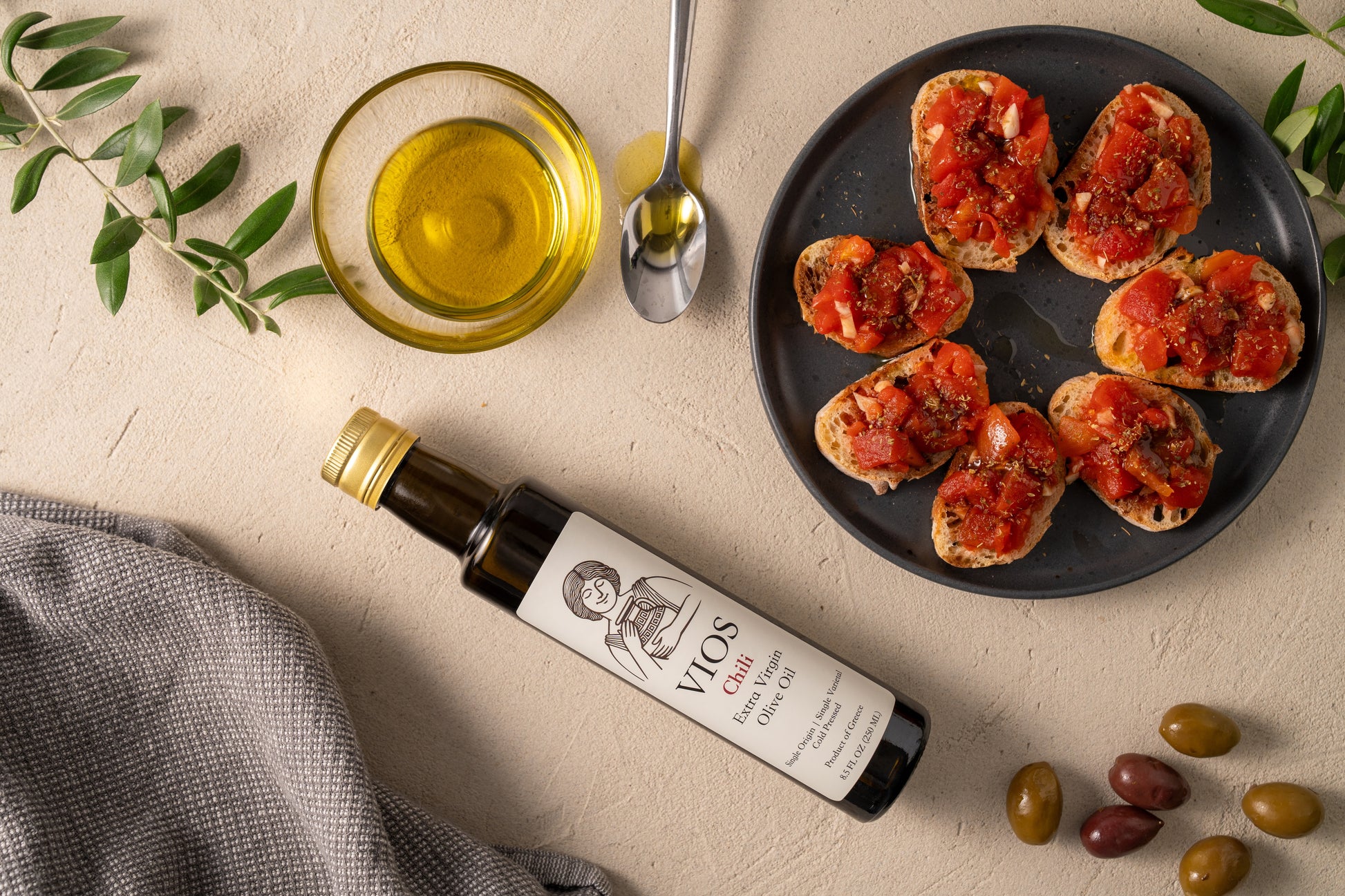 Bottle of VIOS chili extra virgin olive oil with a plate of bruschetta, olives, and bowl of olive oil on a table.