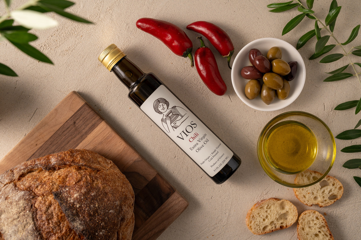 Bottle of VIOS chili extra virgin olive oil with bread, olives, red chili peppers, and a bowl of olive oil on a table.