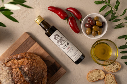 Bottle of VIOS chili extra virgin olive oil with bread, olives, red chili peppers, and a bowl of olive oil on a table.