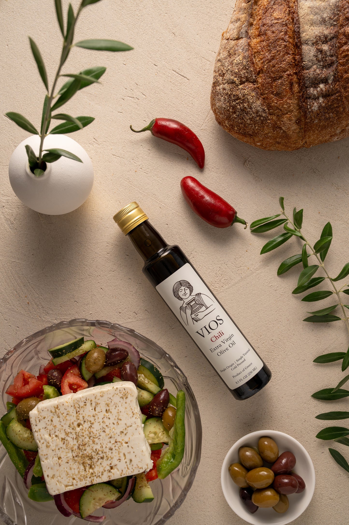 Bottle of VIOS chili extra virgin olive oil with a salad, bread, chilis, olives on a table.