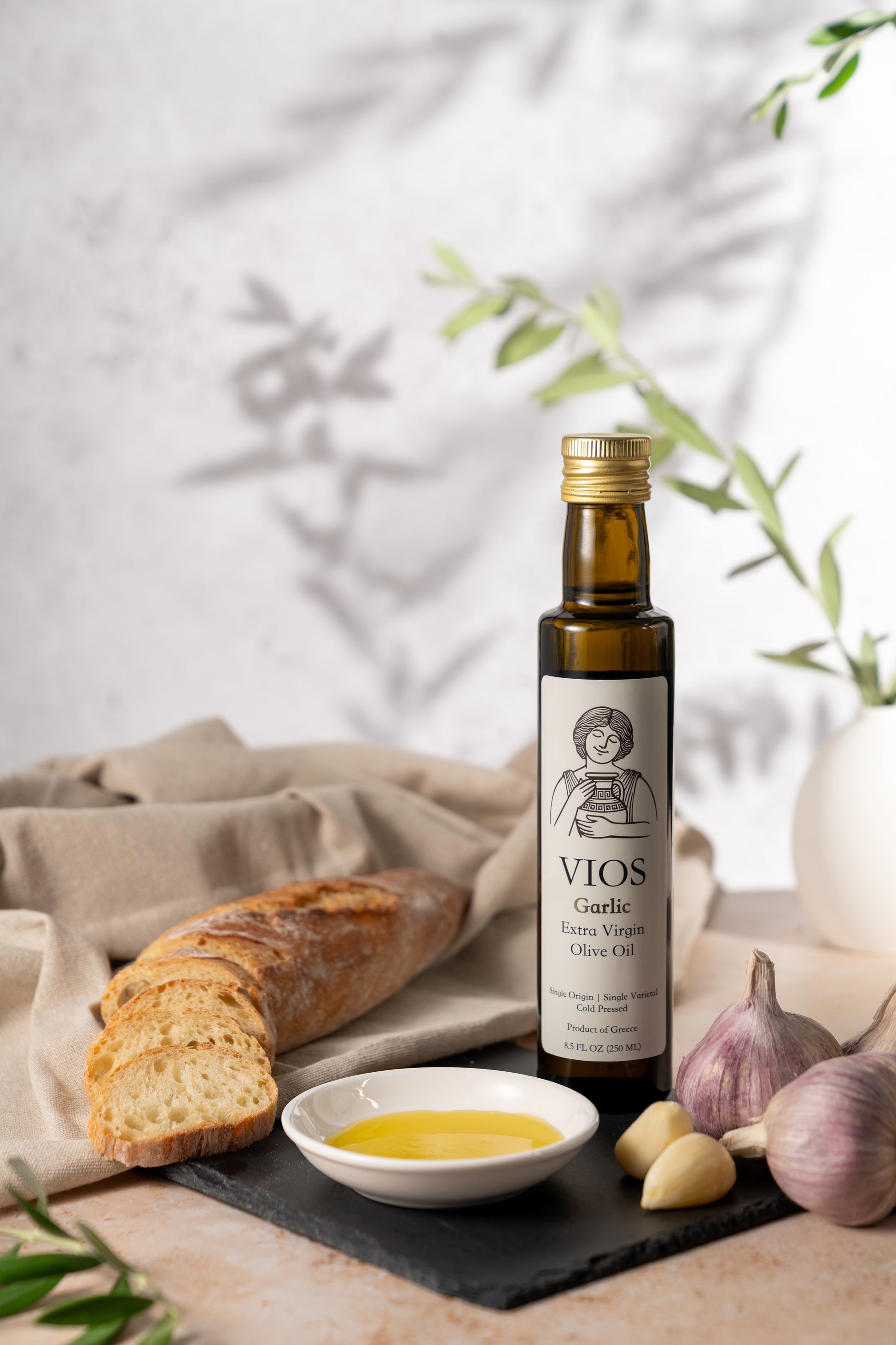 Bottle of VIOS garlic extra virgin olive oil with bread, garlic, and a saucer filled with olive oil on a table