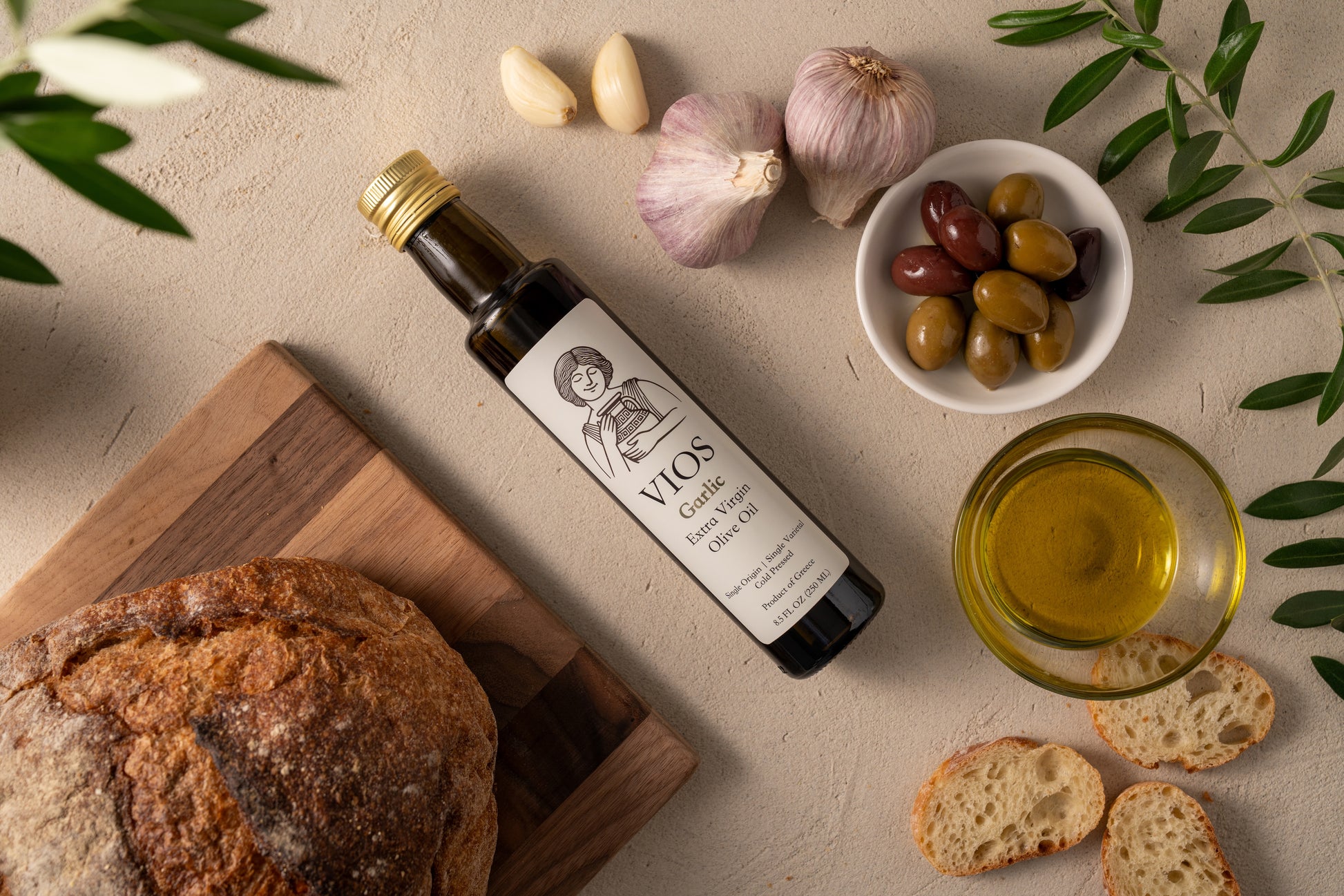 Bottle of VIOS garlic extra virgin olive oil with bread, olives, garlic, and a bowl of olive oil on a table