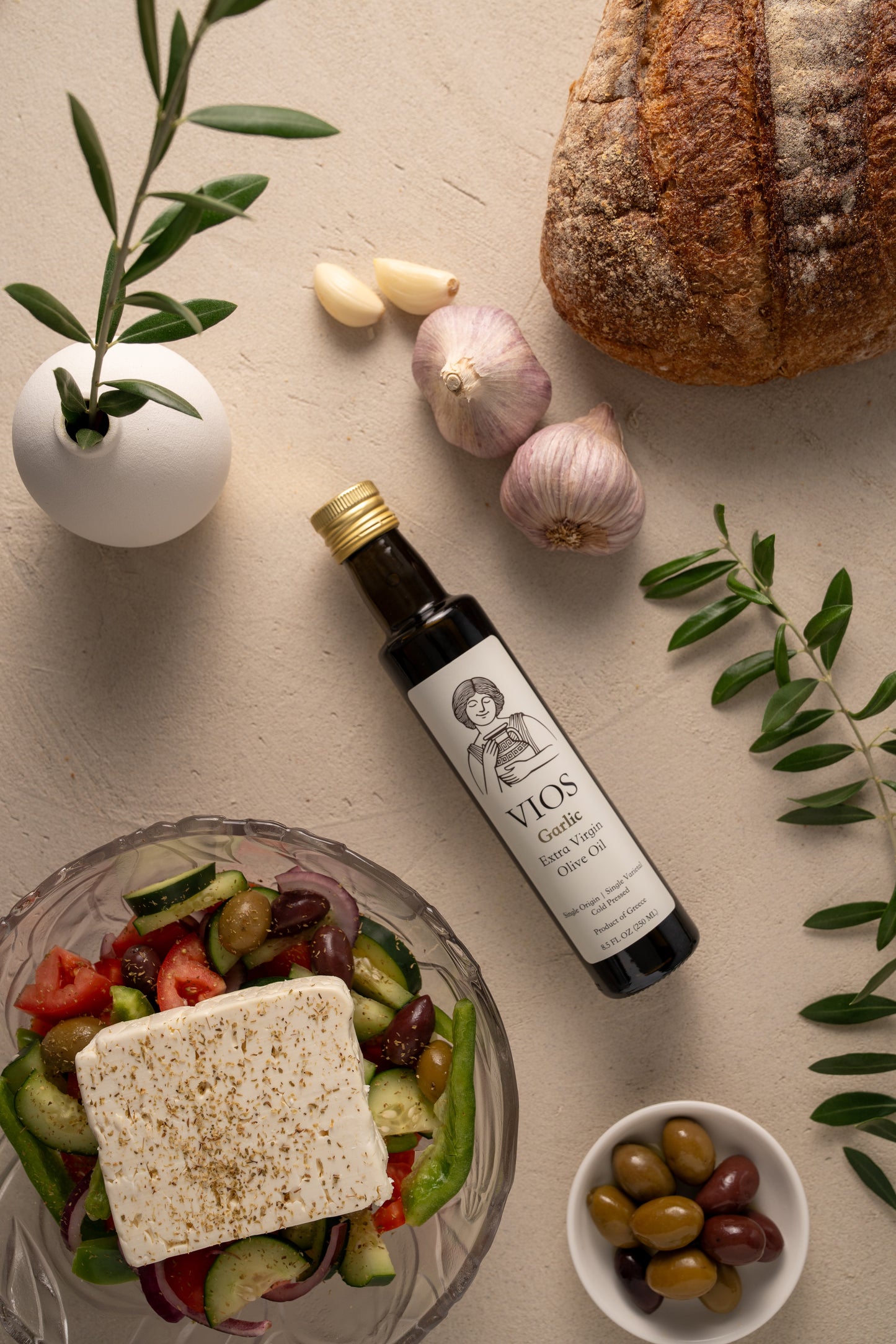 Bottle of VIOS garlic extra virgin olive oil with a salad, bread, garlic, and olives on a table.