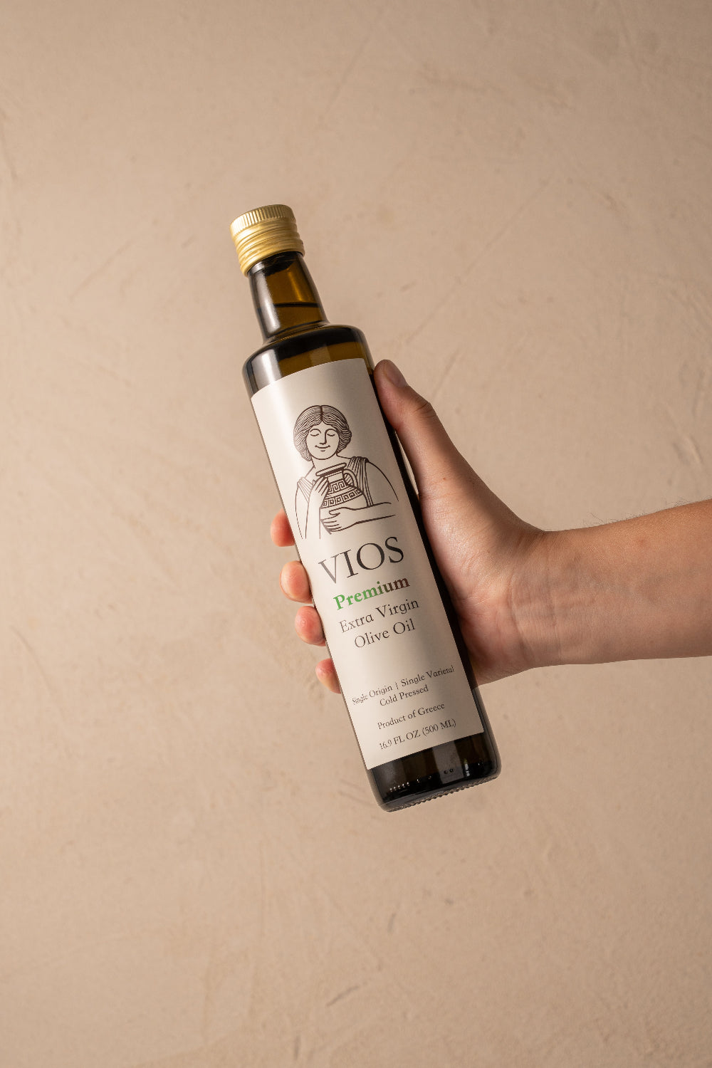 Hand holding a bottle of Vios Premium Extra Virgin Olive Oil against a beige background