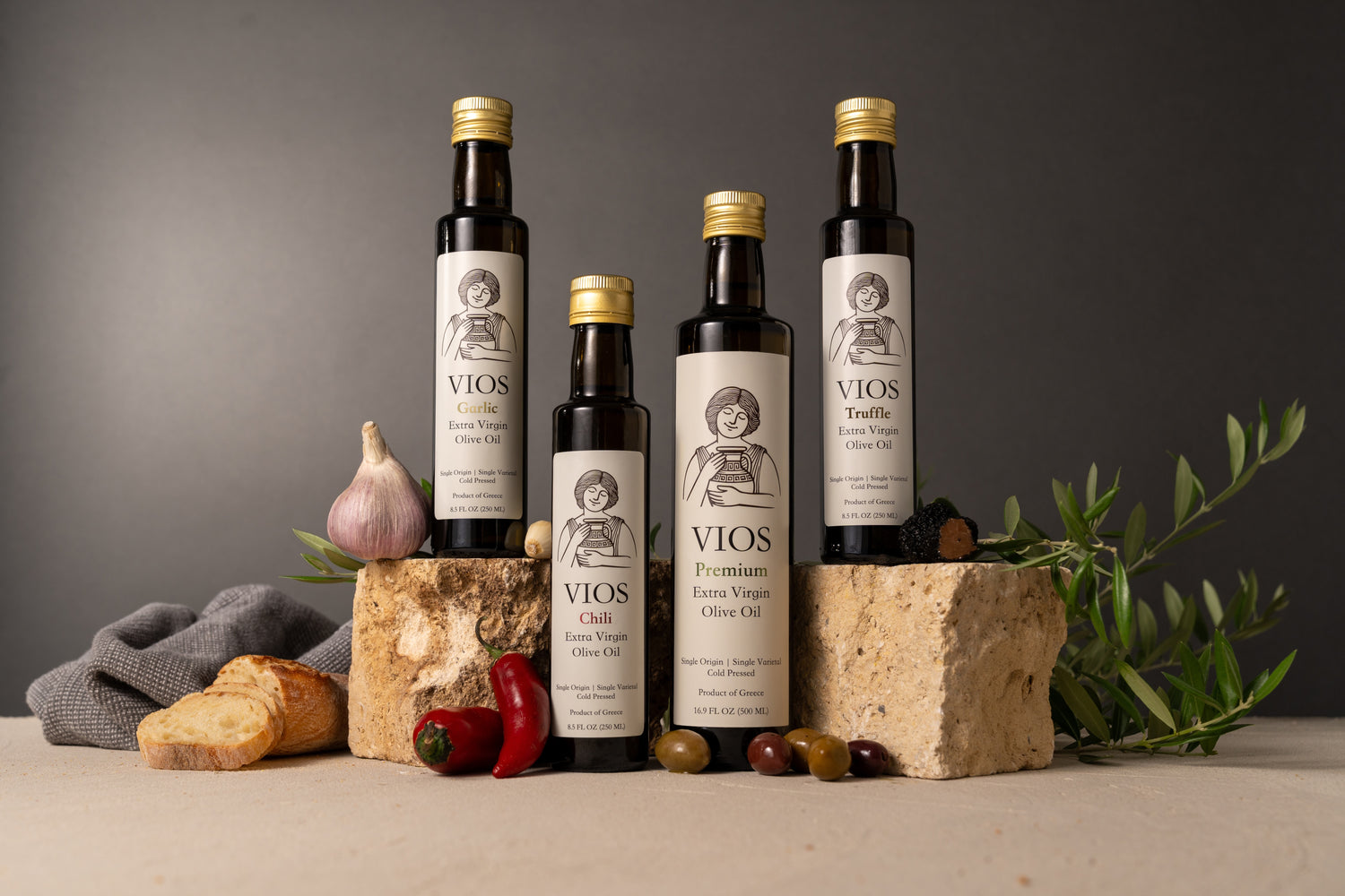 Group shot of 4 bottles of extra virgin olive oil (garlic, chili, premium, truffle) with bread, garlic, red chili peppers, olives, and truffles on stone blocks