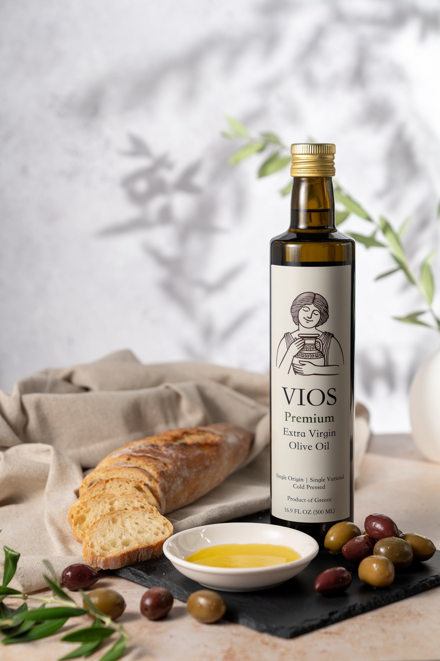 Bottle of VIOS premium extra virgin olive oil with bread, olives, and a saucer of olive oil on a table