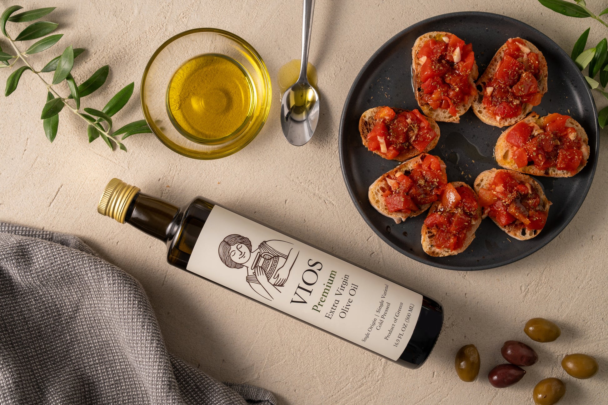 Bottle of VIOS premium extra virgin olive oil with a plate of bruschetta, olives, and a bowl of olive oil on a beige surface