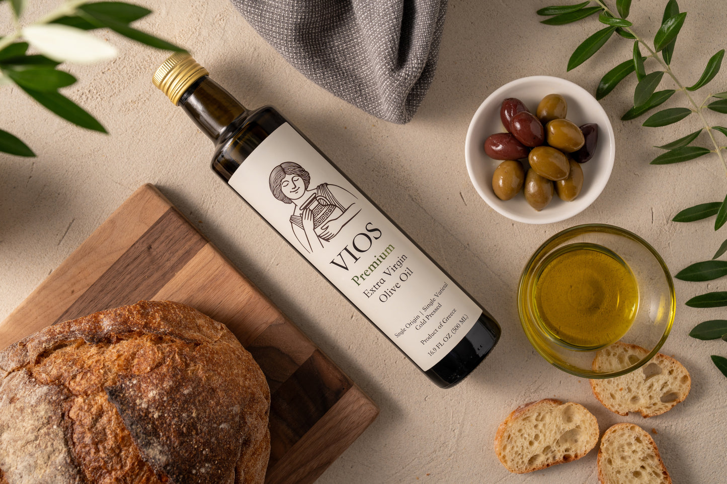 Bottle of VIOS premium extra virgin olive oil with bread, olives, and a bowl of olive oil on a neutral background